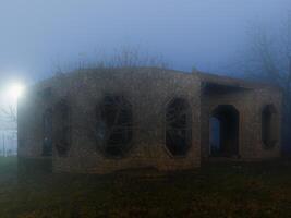 An eerie, abandoned stone building with empty windows stands shrouded in thick fog at night, creating a mysterious and haunting atmosphere. photo
