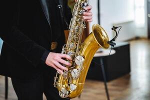 A man dressed in a stylish formal suit skillfully plays a saxophone while standing next to a microphone, producing a captivating melody that beautifully resonates throughout the entire room photo