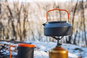 A kettle made of metal and a ceramic cup are resting on top of a gas stove situated in a snowy outdoor setting, surrounded by a blanket of white snowflakes in the chilly air photo