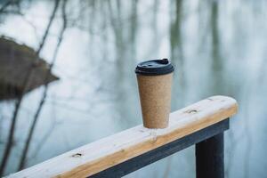 A steaming cup of aromatic coffee is comfortably resting on a rustic wooden railing that is situated right next to a tranquil body of water, creating a serene atmosphere photo