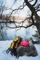 A beautifully colorful backpack is gently resting on a snowcovered rock, right by a tranquil lake, situated in a magical winter wonderland, adorned with glittering snow and sparkling ice photo