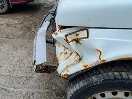 In a parking lot, there is a white car that has a visibly damaged bumper, indicating that it may have been involved in an accident or traffic collision, and is now parked there photo