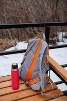 A colorful backpack and a shiny thermos are sitting side by side on a wellworn wooden table, creating a cozy and inviting scene. Both items hint at an adventure awaiting them photo