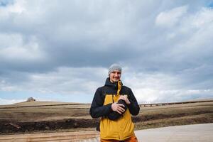 A man in a bright yellow jacket stands confidently in an expansive field, skillfully using his camera to capture the breathtaking beauty of the picturesque landscape around him photo