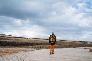 A man, equipped with a sturdy backpack, is strolling leisurely along an extended wooden walkway that gracefully winds through a beautiful and scenic area surrounded by nature photo
