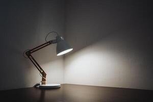 A stylish desk lamp is prominently sitting on a wooden table in an otherwise dark room, casting soft light and creating intriguing shadows that enhance the atmosphere within the space photo