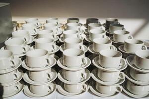 A large collection of white coffee cups and saucers are meticulously stacked on top of each other upon a table, creating an organized and aesthetically pleasing display of serveware photo