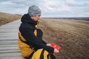 A man wearing a bright yellow and black jacket is sitting comfortably on a wooden walkway, intently looking over a map as he plans his next steps in the beautiful outdoor surroundings photo