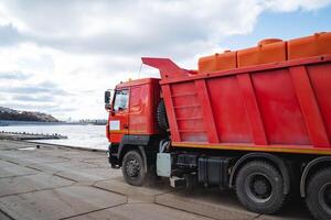 A bright red dump truck is parked alongside a serene body of water, adding a splash of color to the picturesque landscape and showcasing its robust design and functionality photo