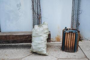 A bag filled with garbage is currently sitting right next to a wooden trash can, which provides an interesting contrast in this still life composition that captures everyday objects photo