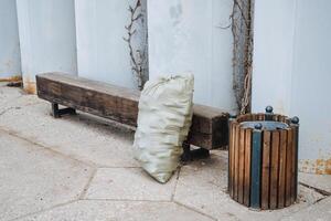 An unappealing bag of trash is currently resting on a sturdy wooden bench that is positioned right next to a similarly made wooden trash can, creating a scene of urban neglect photo