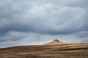 At the summit of a gentle hill, there stands a modest little building, surrounded by a vast expanse of green field that stretches into the horizon beyond the rolling landscape photo