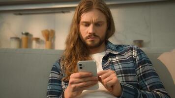 Calm serious Caucasian man guy male user gadget addict indoors at home couch comfortable sofa using mobile phone scrolling social apps media reading watching with smartphone web net e-commerce photo