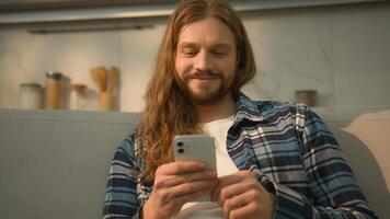 Happy smiling carefree positive relaxed Caucasian man guy male user indoors at home couch comfortable sofa using mobile phone scrolling social apps media laughing reading watching with smartphone web photo