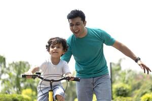 Cute Boy Learning To Ride A Bicycle In A Park With The Help Of His Father photo