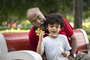 Happy Kid Blowing Soap Bubbles In Park With Grandfather While Sitting On Bench In Park photo