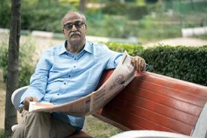 Old Man Sitting In Chair In Park While Reading Newspaper photo