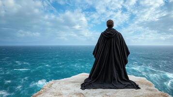 A person in a black cloak standing on a cliff overlooking the ocean photo