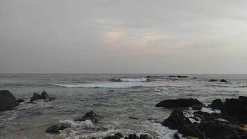 calm ocean scene with waves breaking over dark coastal rocks under a soft, overcast sky. The muted tones evoke a serene and peaceful coastal vibe, ideal for nature or travel concepts video