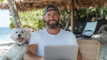 A man with a beard and a dog sitting on a beach with a laptop photo