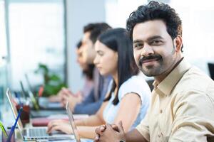 Excited Team Member In Cream Shirt Sitting At Desk Smiling Looking At Camera photo