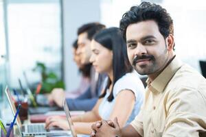 Work Colleague In Shirt Working On Laptop While Looking At Camera In Office photo
