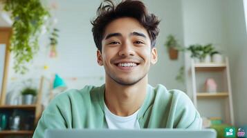 A young smiling man sitting in a comfortable home environment teaching a virtual class on his laptop while surrounded by bookshelves and greenery photo