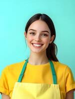Cheerful and confident young woman wearing a yellow shirt and apron smiling directly at the against a vibrant turquoise background photo