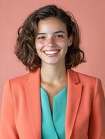 Close up portrait of a confident friendly young woman with curly brown hair and a warm smile dressed in a stylish coral blazer and mint green blouse against a vibrant orange background photo