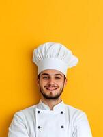 Headshot portrait of a confident bearded young chef wearing a traditional white uniform and hat smiling in front of a vivid yellow studio background photo