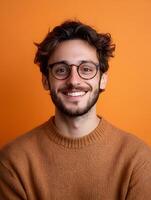 Portrait of a friendly smiling young man with glasses and a beard wearing a warm brown sweater against a vibrant orange background photo