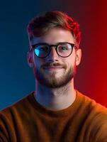Portrait of a young smiling man with glasses and a beard wearing a casual orange sweater against a colorful blue and red background suggesting a creative photo