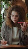 Vertical . Freelance employee talking on smartphone using speaker mode, taking notes sitting at desk. video
