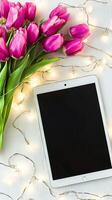 Pink Tulips and Digital Tablet on a White Surface with String Lights. Concept of Spring Decor, Modern Workspace, Technology in Nature. Mockup. Copy space photo