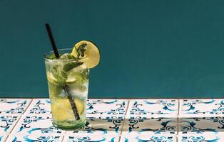 Close-up of a glass of delicious mojito with lime and fresh mint. Tasty lemonade with ice cubes on a table with patterned tiles. A cup of cool citrus cocktail and a straw. photo