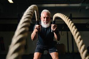 Senior man demonstrates strength and fitness, intensely working out with battle ropes at the gym. photo