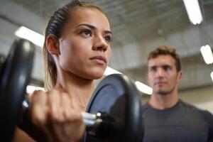 Focused woman lifts dumbbell with personal trainer watching her form in a modern gym. photo