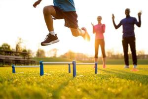 Young athlete leaps over a hurdle on a sunny field with parents cheering him on during training. photo