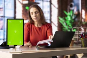 Person at her home office reading news and research papers with green screen, virtual connection online. Woman collecting notes and data from different sources to know current events. photo