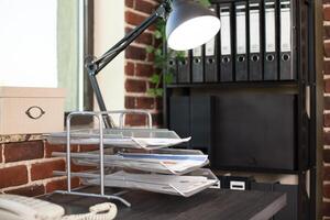 Lamp, documents and telephone arranged on wooden desk with bookshelf set nearby in brick wall room. Closeup of empty workspace with table, office cabinet, and paperwork well organized for work. photo