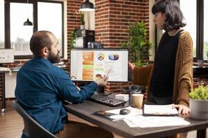 Businessman pointing at computer screen and updating colleague on company market analysis in startup office. Caucasian woman listening to male coworker explain project presentation on desktop pc. photo