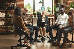 Senior woman psychologist advising support group of anxious patients at mental health rehabilitation meeting. Old white man shares his journey of recovery in multiethnic circle during therapy session. photo
