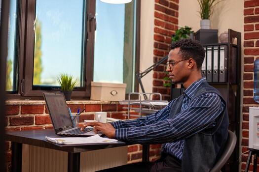 Office worker researching company data on laptop, analyzing startup information for business growth. Black man sitting at desk and using personal computer to gather insights for project development. photo