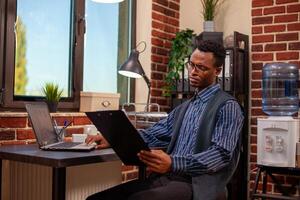 Black man manager sitting at desk and reading documents with a focused expression. Busy analyst with glasses, holds clipboard, reviewing information and comparing with research data on laptop. photo