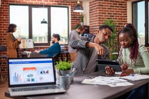 Black employees examine company data on tablet, brainstorming ideas in startup office. Male and female coworkers collaborate on strategy development and reviewing business reports on digital device. photo