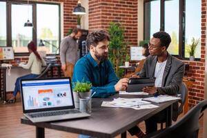 Male entrepreneurs brainstorming business ideas and reviewing company data on a tablet in modern workplace. Young men seated at desk with shared device, analyzing annual reports collaboratively. photo