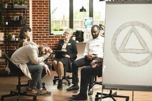 Group of alcoholics gather in aa meeting room with symbol displayed on white board. Retired therapist conducting supportive conversation, discussing mental health and addiction in brick wall office. photo