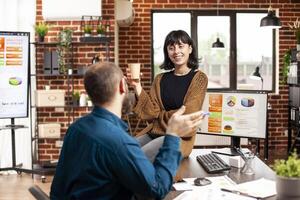 Smiling businesswoman with coffee, sitting on office desk and discussing with male coworker in brick wall room. Caucasian colleagues taking a break and having conversation in startup office. photo