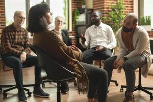 Young and retired individuals sitting in brick wall room and having discussion about mental health at therapy session. Diverse group listening to white woman speak about her feelings during aa meeting photo
