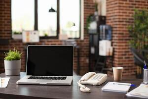 Startup office desk arranged with plant, phone, notebook and digital device displaying blank screen. Vacant table set with open laptop, documents and coffee, creating ready for work atmosphere. photo